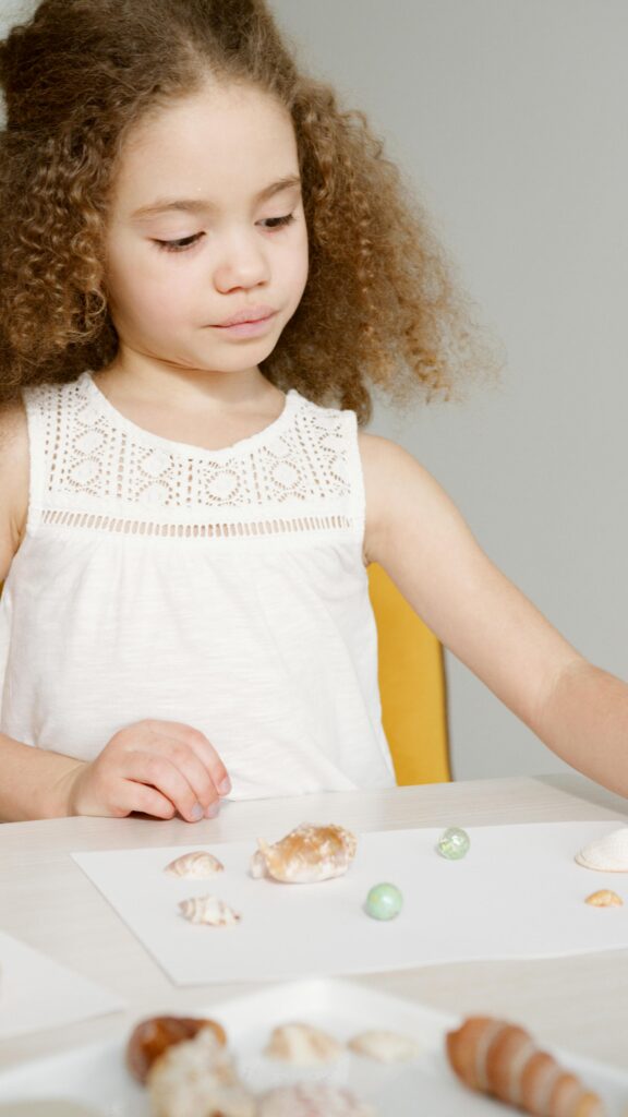 Young girl with curly hair arranging shells on paper, learning through play.