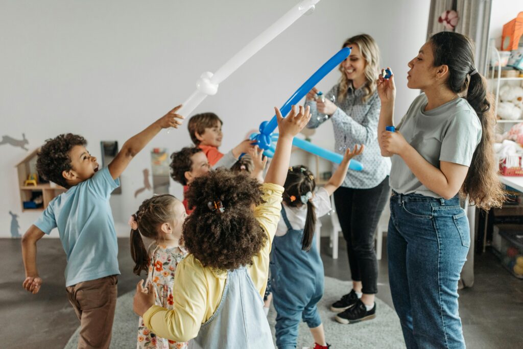 Group of children and teachers enjoying balloon games indoors in a kindergarten setting.